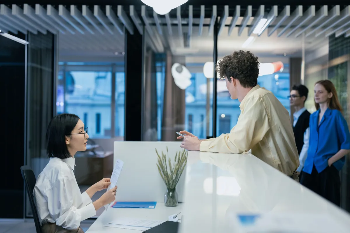 A receptionist helps a visitor at the front desk while others wait